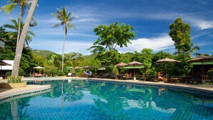 Une piscine d’un bleu éclatant, bordée de carreaux, entourée de chaises longues et de parasols, de palmiers et dominée par un paysage montagneux.