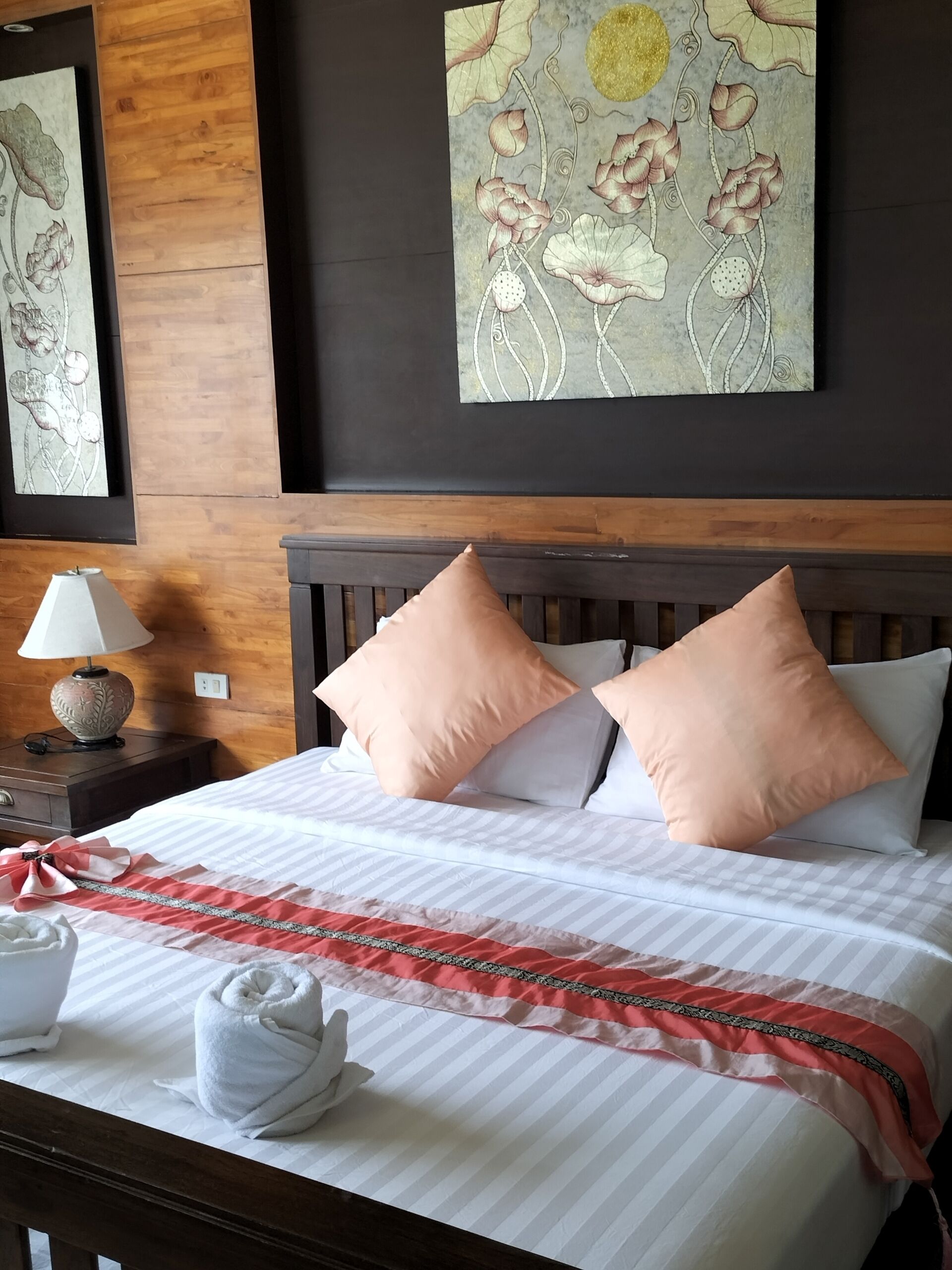 Una cama bien hecha con una colcha a rayas y un borde decorativo rojo y negro, dos almohadas, una toalla blanca y una mesita de noche con una lámpara.