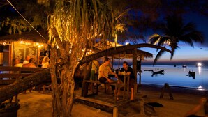 Une construction en bois située au bord de la plage, dotée d’un toit de chaume, entourée de palmiers et offrant une vue sur des eaux calmes.