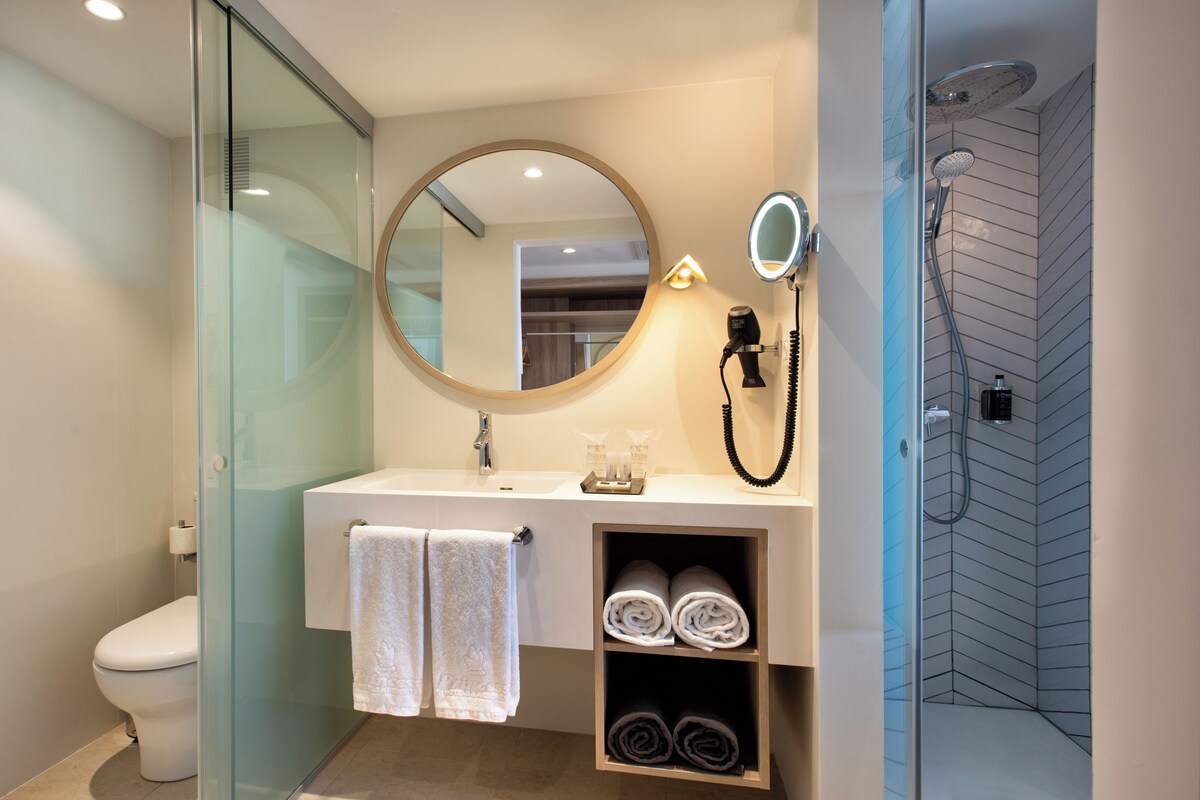 Une salle de bain moderne avec une douche à parois de verre, un miroir rond, un sèche-cheveux et des serviettes soigneusement pliées.