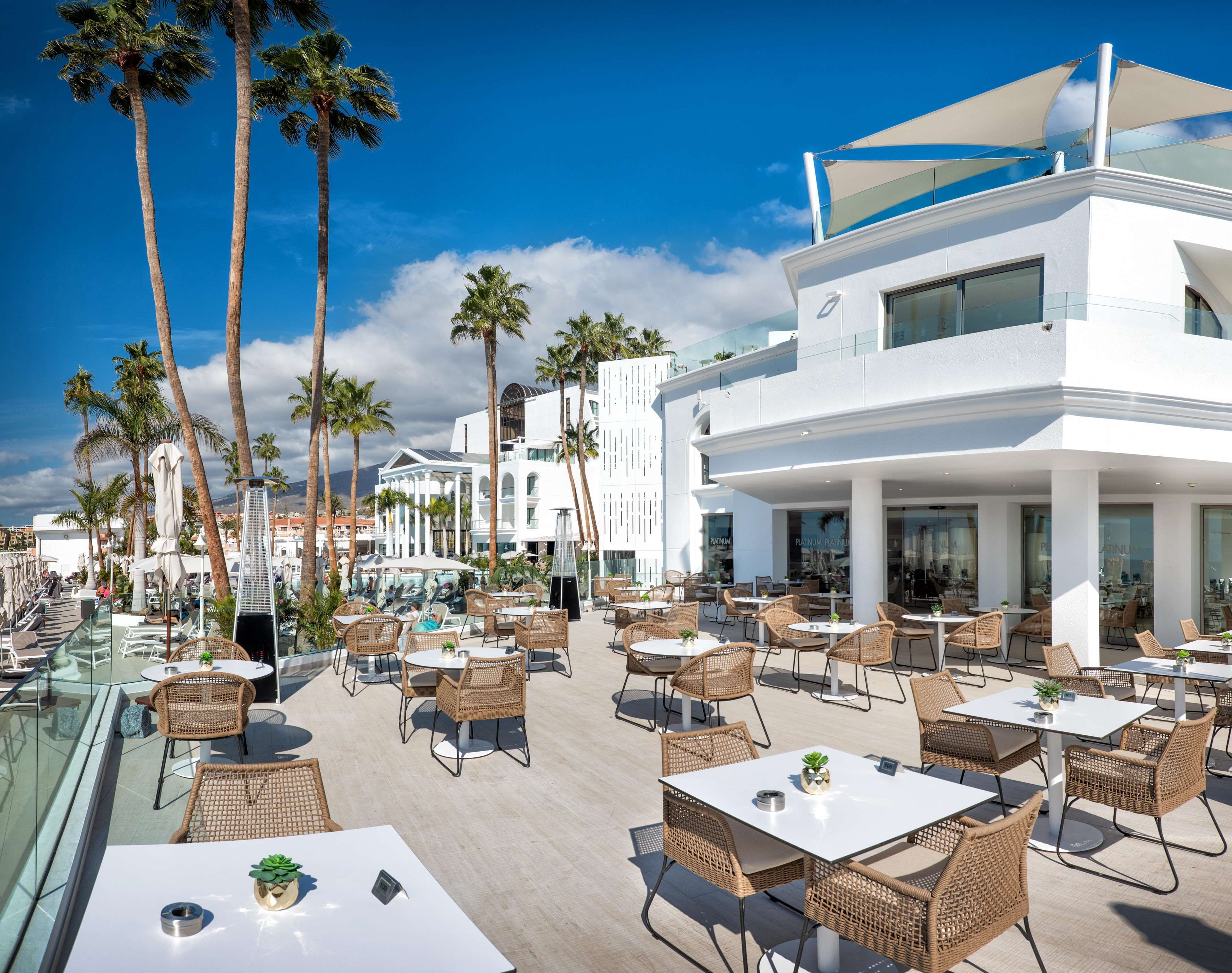 An outdoor dining area with wicker furniture and white tables, surrounded by palm trees and a modern building with large windows.