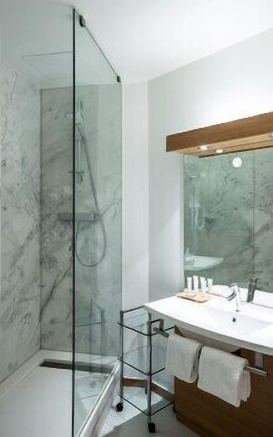Un baño con cabina de ducha de vidrio, un lavamanos blanco sobre un mueble de madera y pared de mármol.