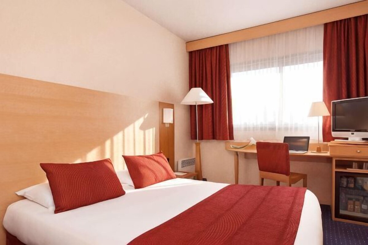 A hotel room with a bed, desk, chair, TV, and window with curtains.