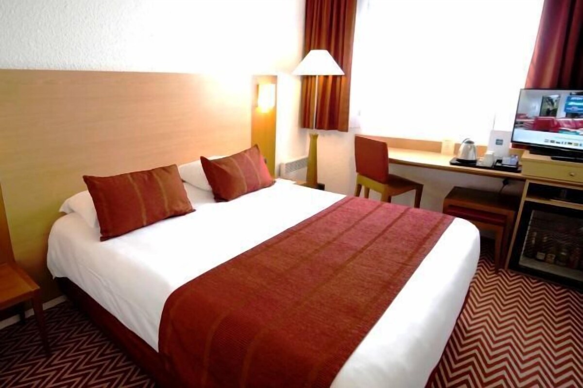 A hotel room with a bed, a desk, a chair, a lamp, and a television.