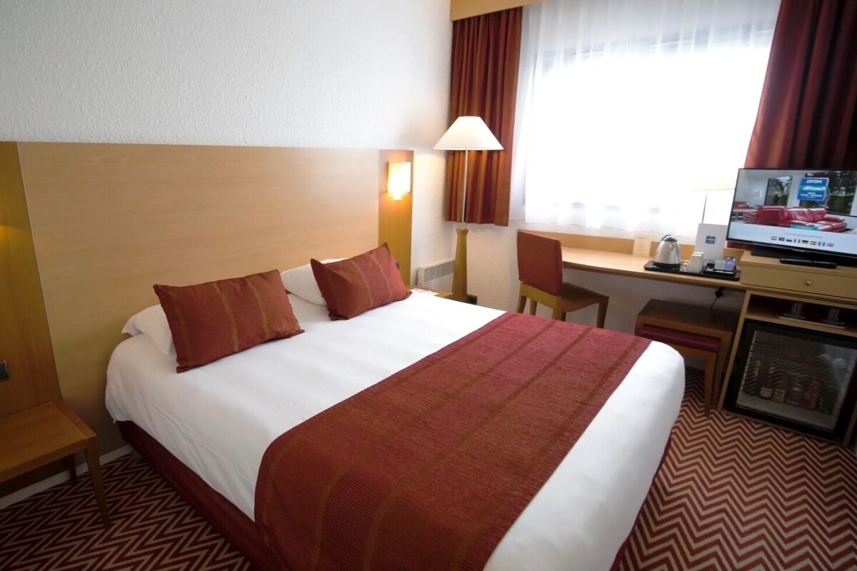 A hotel room with a bed, desk, chair, TV, and a minibar.