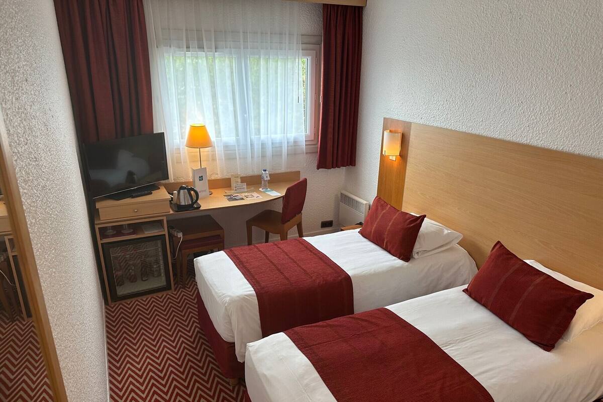 Ein Hotelzimmer mit zwei Betten, einem Schreibtisch mit Computer, einem Fernseher und einem Fenster mit Vorhängen.