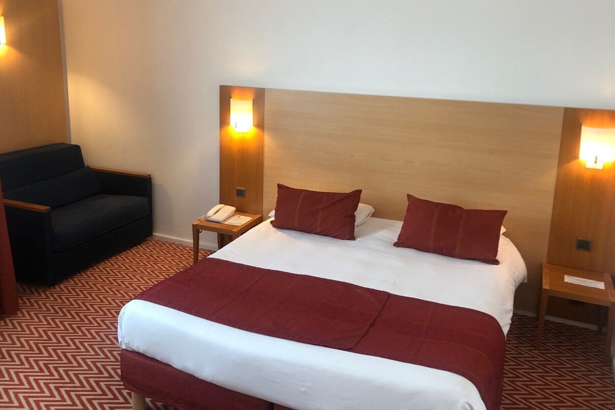 A hotel room with a large bed, two bedside tables, a chair, and two wall-mounted lamps.
