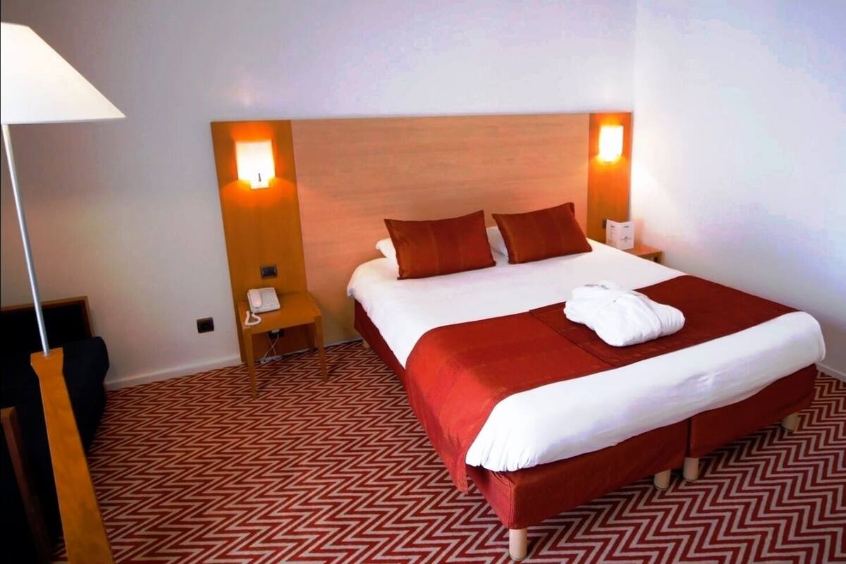 Ein Hotelzimmer mit einem großen Bett, zwei Nachttischlampen, einem kleinen Nachttisch mit Telefon und einem Spiegel an der Wand.