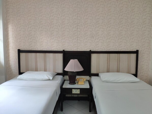 Habitación de hotel con dos camas individuales, una mesita de noche con lámpara y teléfono, y un papel tapiz con motivos florales.