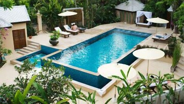 Una piscina rectangular con borde de azulejos, rodeada de sillones de descanso y sombrillas, ubicada en un jardín exuberante con diversas plantas y un edificio pequeño al fondo.