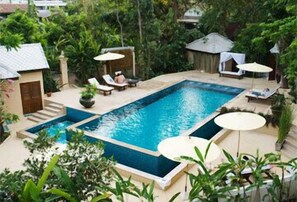 Una piscina rectangular con borde de azulejos, rodeada de sillones de descanso y sombrillas, ubicada en un jardín exuberante con diversas plantas y un edificio pequeño al fondo.