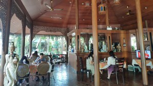 Un área de comedor cubierta con techo y columnas de madera, mesas con sillas y elementos decorativos.
