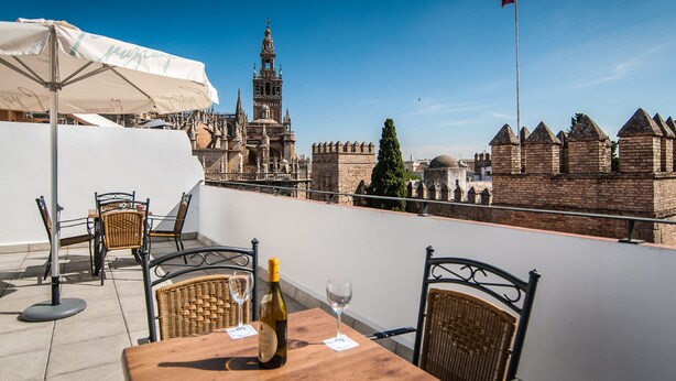 Eine Terrasse mit Tisch, Stühlen und einer Weinflasche, mit Blick auf historische Gebäude und eine Flagge.