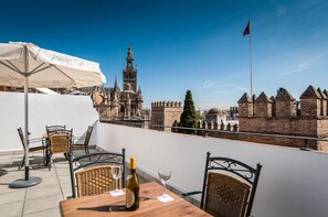 En terrasse med et bord, stole og en vinflaske, med udsigt over historiske bygninger og et flag.