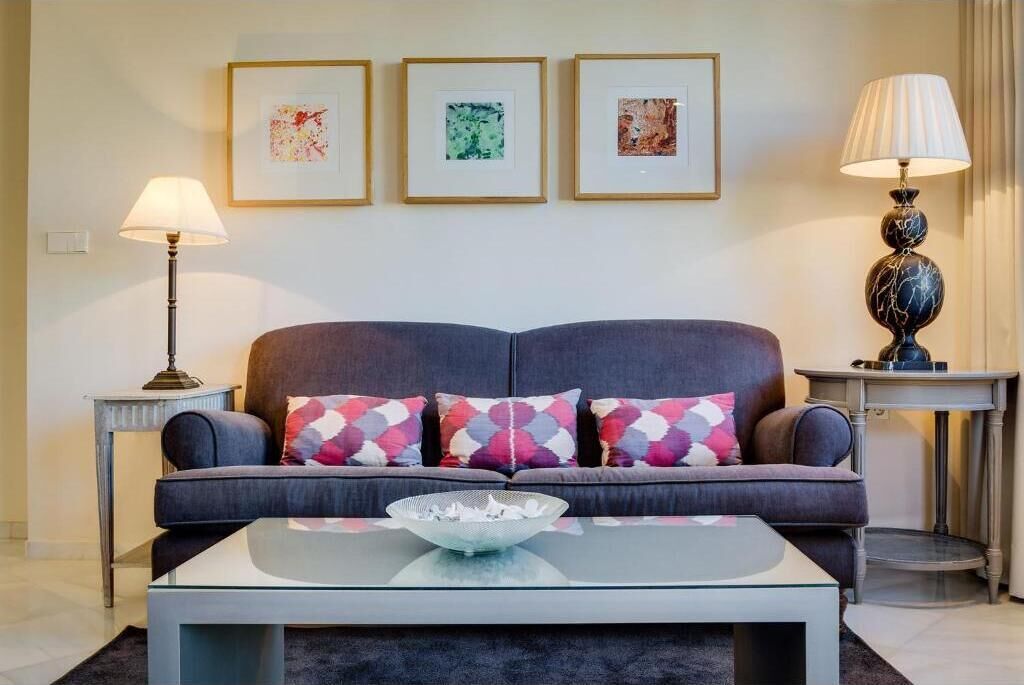 Una sala de estar moderna con un sofá gris, cojines coloridos, una mesa de centro de vidrio y obras de arte enmarcadas en la pared.
