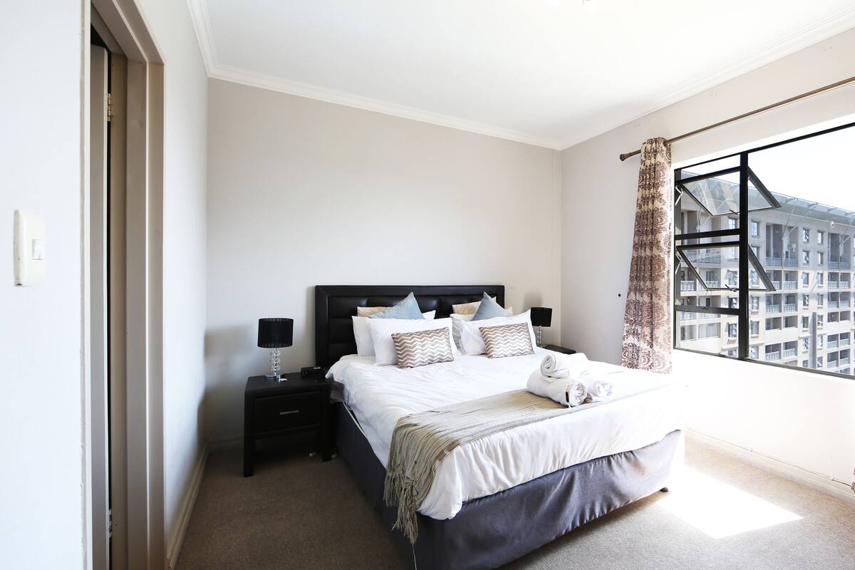 A bedroom with a large bed, a nightstand, a lamp, and a view of a building through a window.