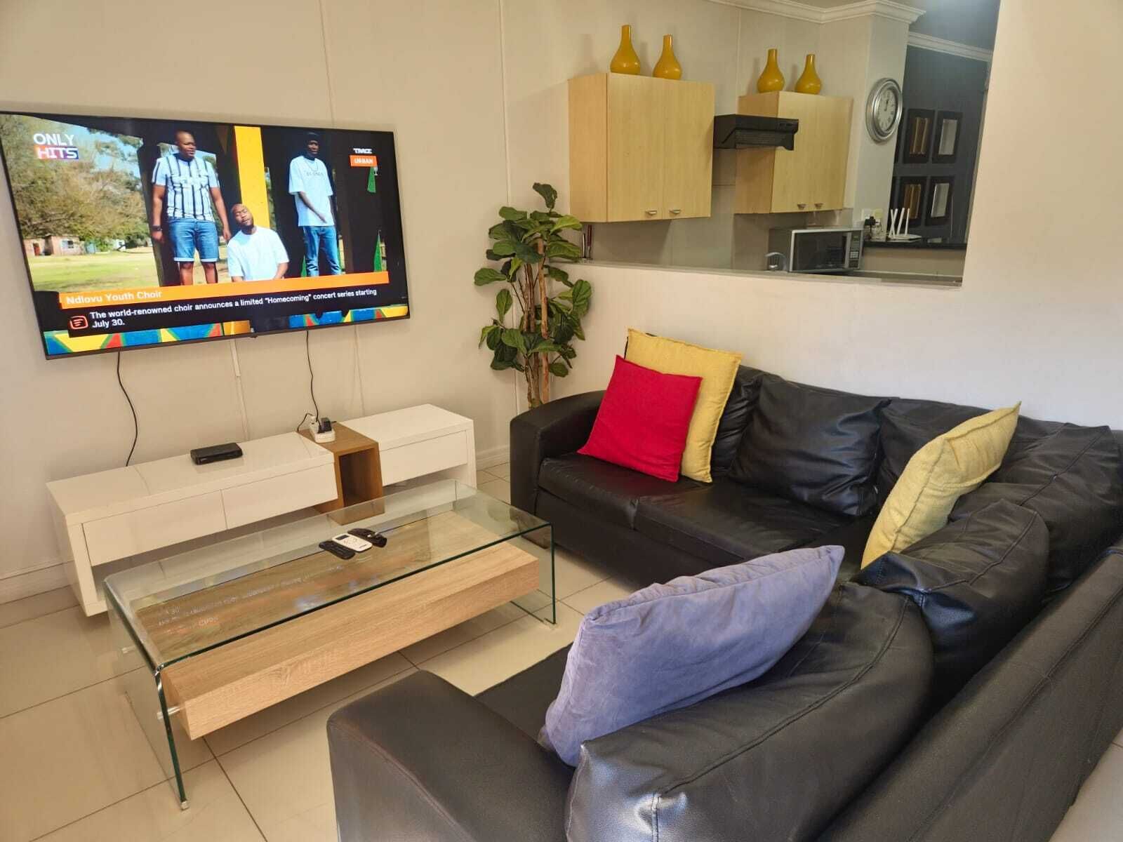 Una sala de estar con un televisor de pantalla plana montado en la pared, un sofá de cuero negro con cojines, una mesa de centro de vidrio y un mueble blanco.