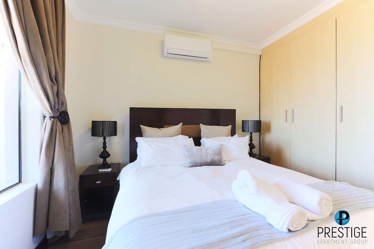 A hotel room with a large bed, two bedside tables with lamps, an air conditioning unit, and a wardrobe.