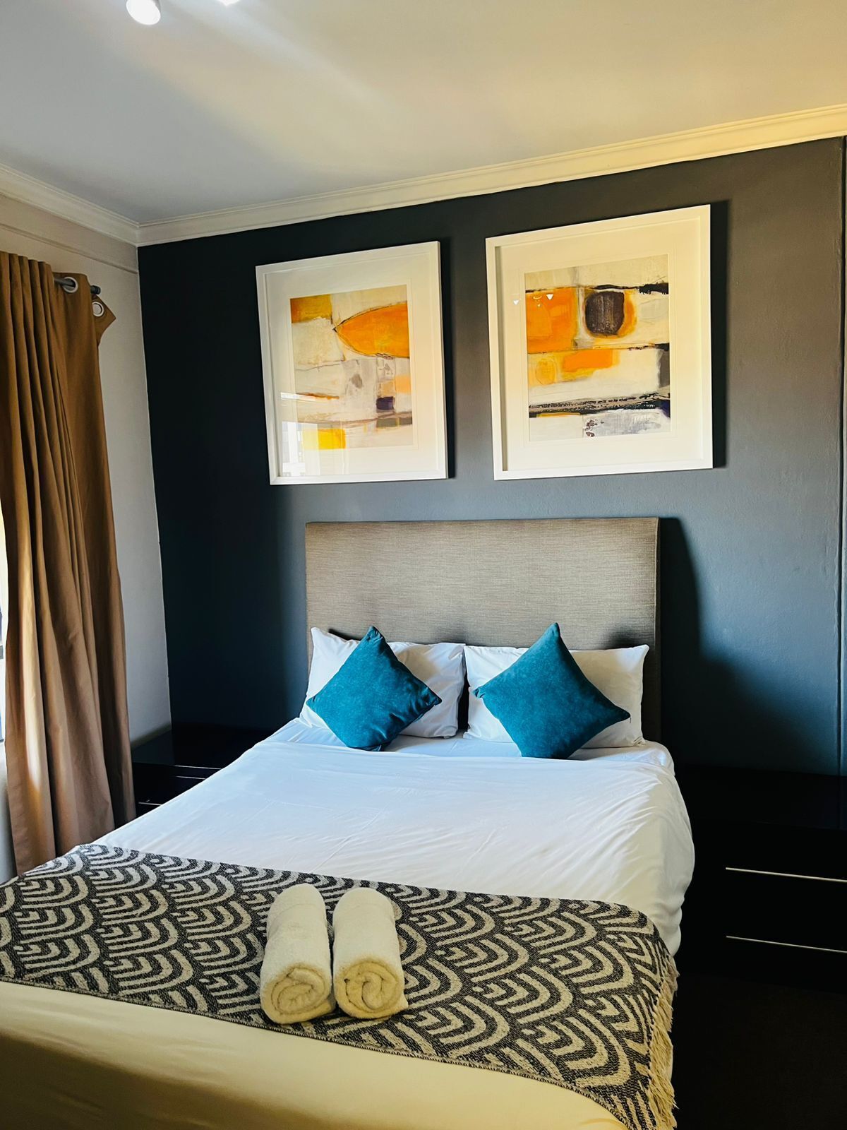 A hotel room with a bed, two framed abstract paintings, a headboard, and a patterned throw blanket.