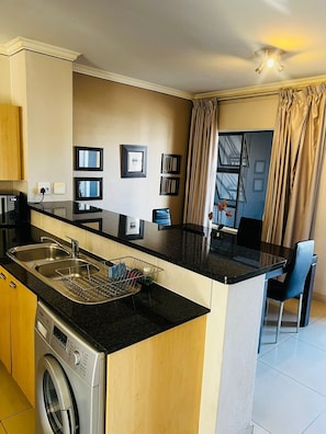 A compact kitchen with a built-in washing machine, a dining area with a table and chairs, and a window with curtains.