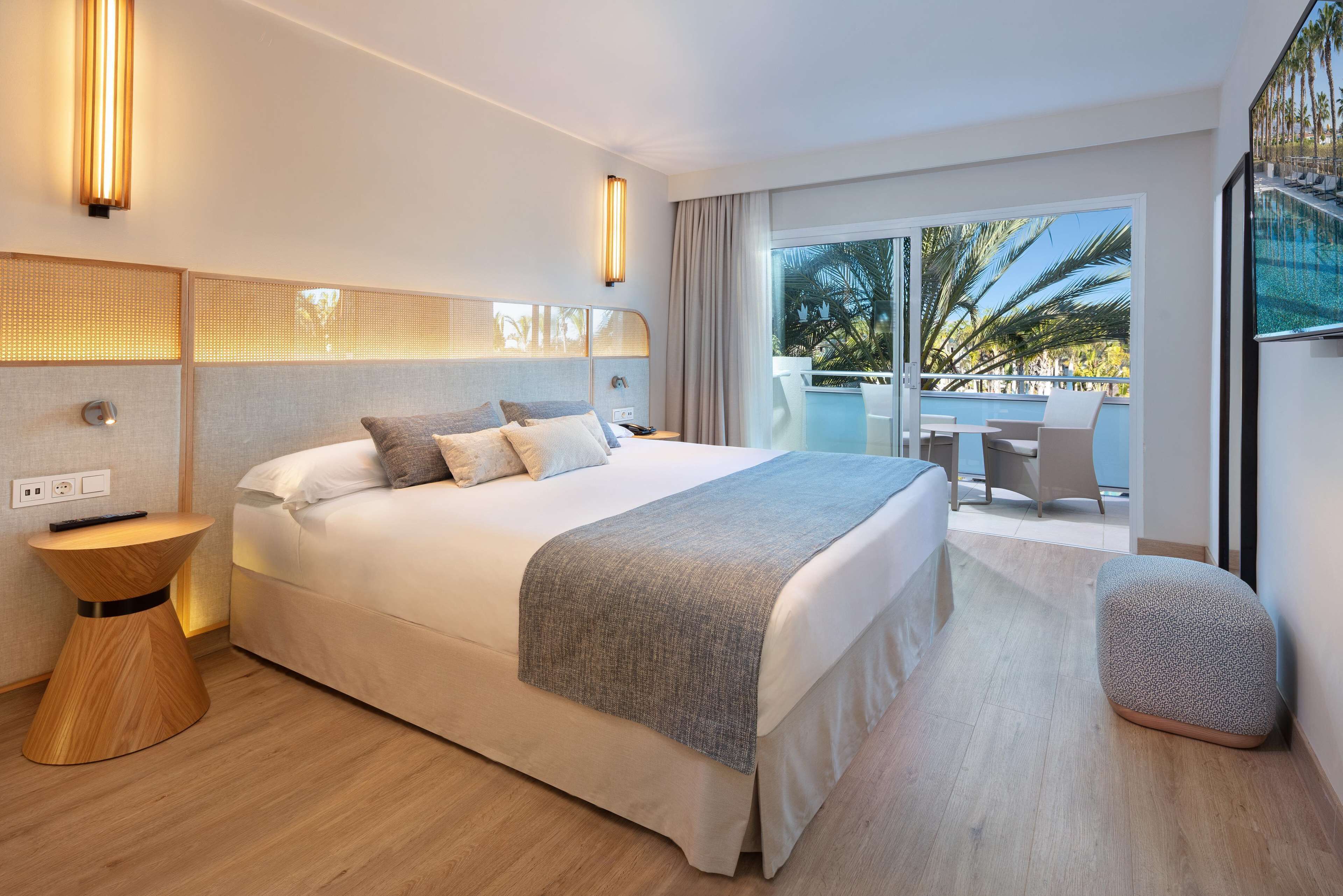 A modern hotel room with a large bed, a side table, a small ottoman, and a balcony with a pool view.