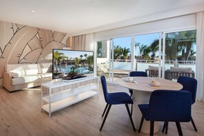 A modern living room with a flat-screen TV, a sofa, a dining table with chairs, and a large window with a view of a pool and palm trees. - Hotel Tabaiba Princess- All Inclusive (San Bartolomé de Tirajana)