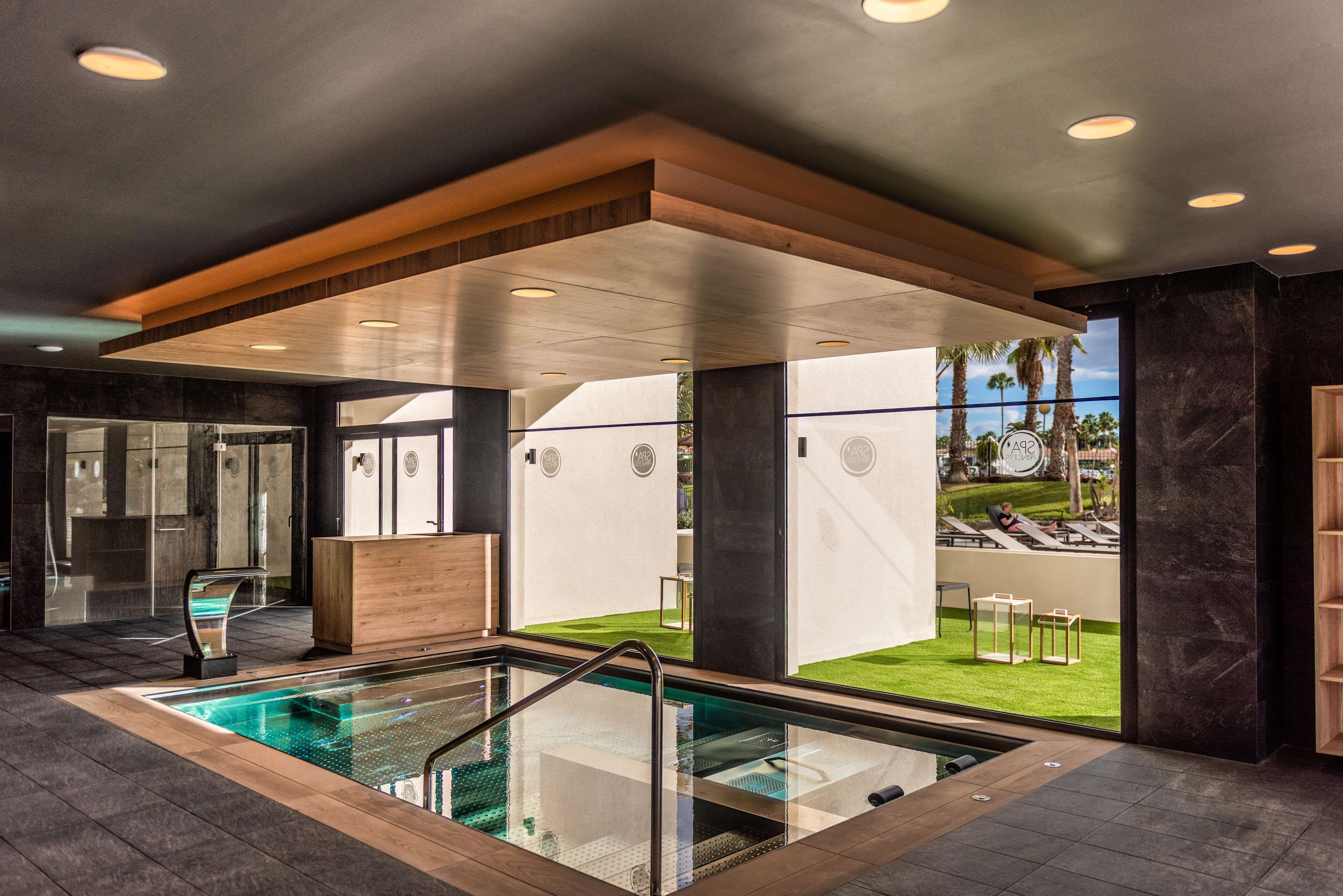 Un área de piscina interior moderna con cerramiento de vidrio, vigas de techo de madera y vistas a un jardín exterior con sillones de descanso.