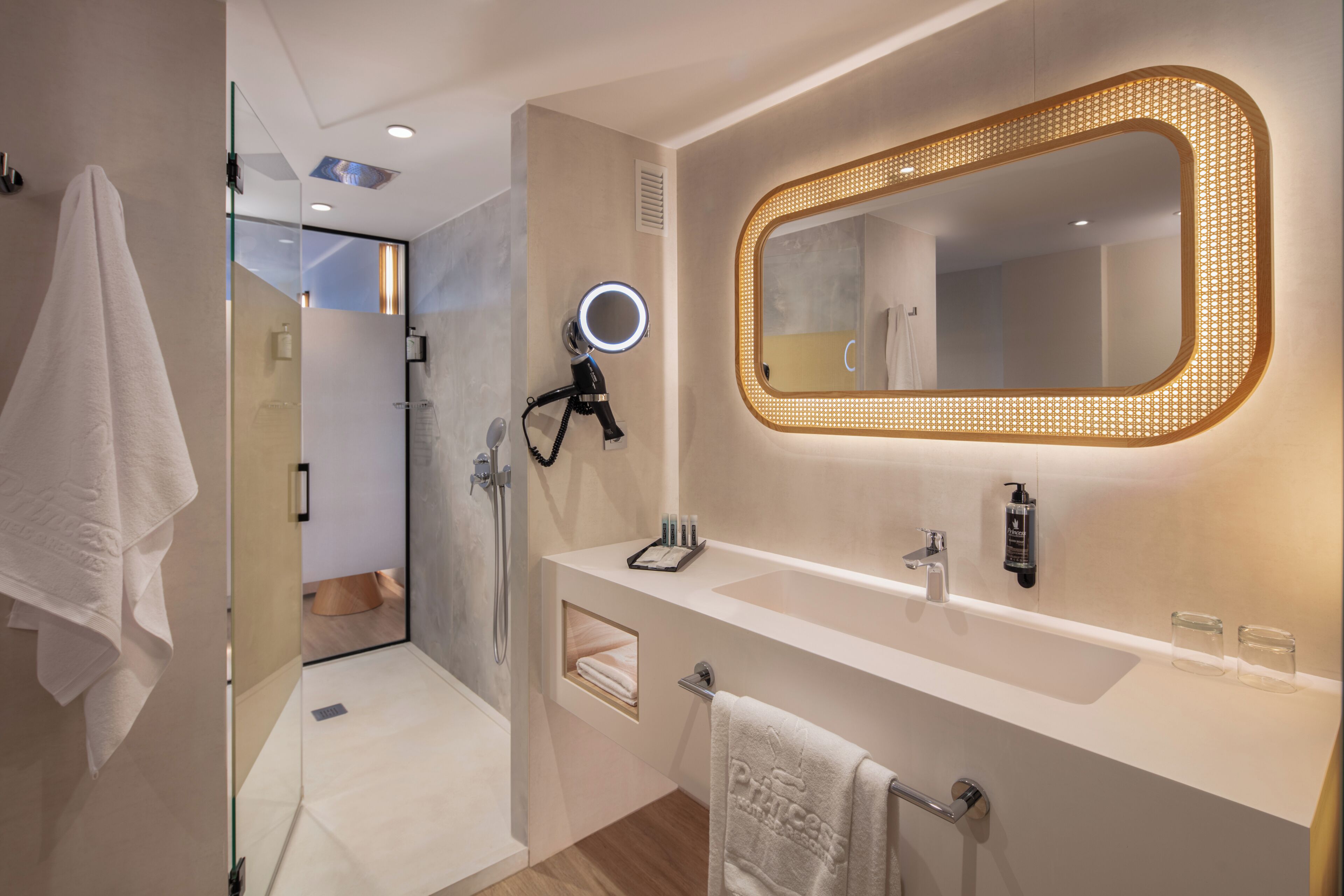 A modern bathroom with a vanity, a large mirror with lights, a hairdryer, and a towel rack.
