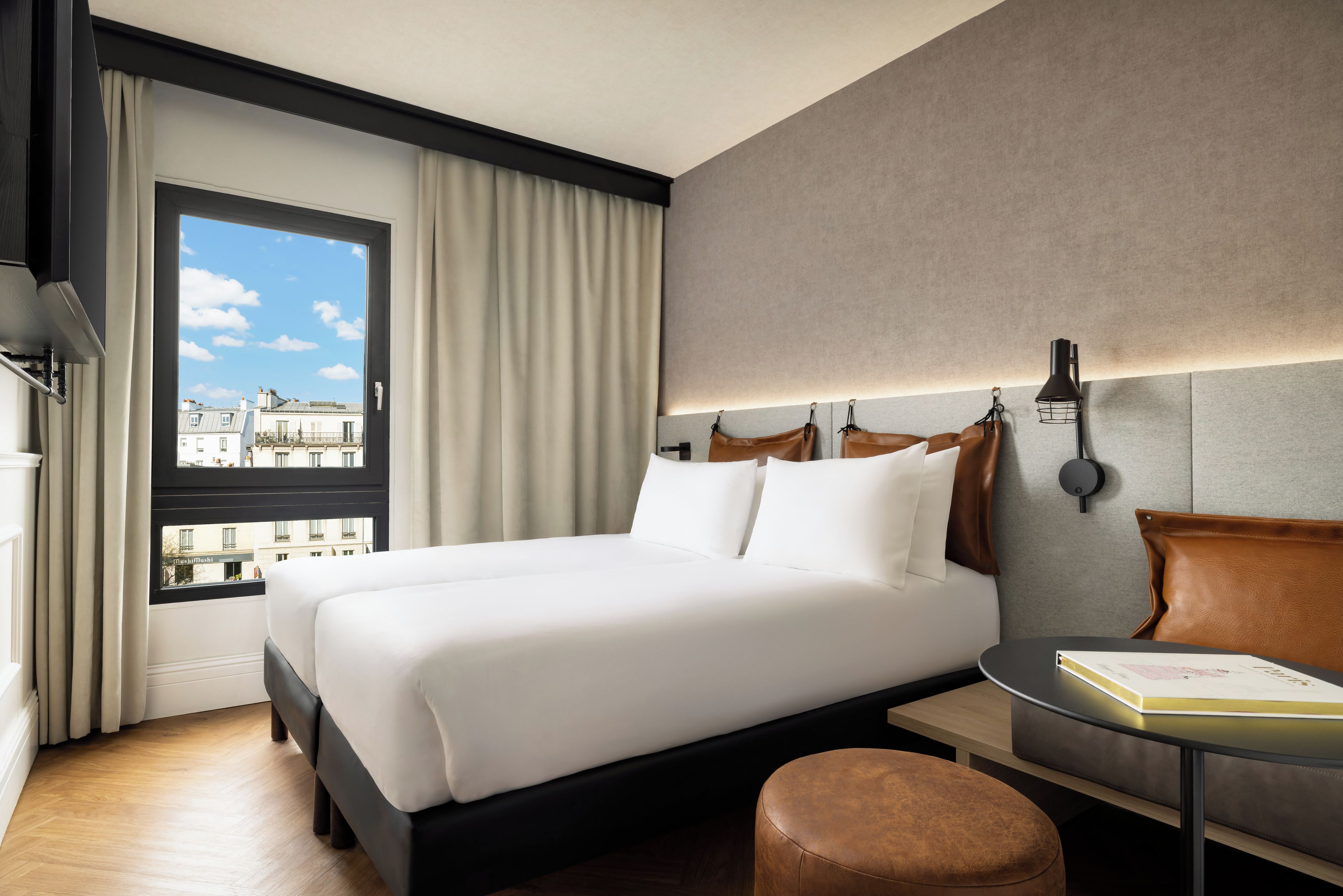 A hotel room with a large bed, a bedside table, a reading lamp, and a view of buildings through the window.