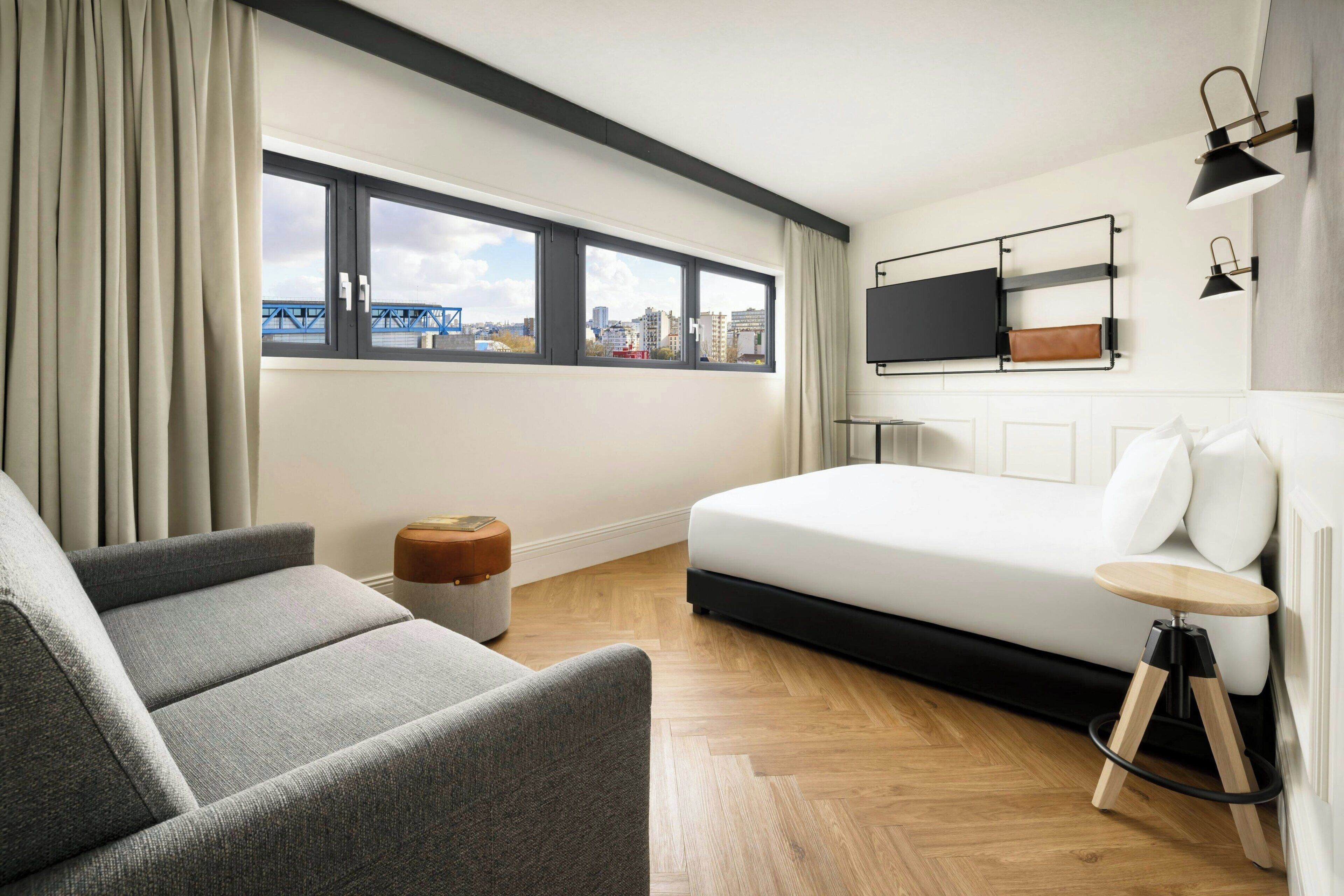 A modern hotel room with a large bed, a sofa, a small stool, a TV mounted on the wall, and a window with a city view.