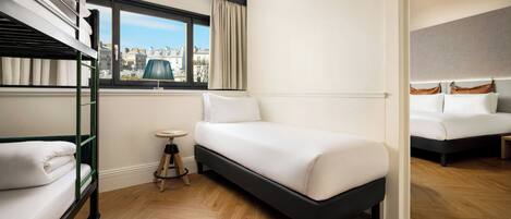 A hotel room with a bunk bed, a single bed, a small round table, and a view of the city through a window.