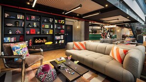 A modern living room with a sofa, armchairs, and a coffee table. There are bookshelves filled with books and decorative items. A staircase is visible in the background. - Moxy Paris La Villette (Paris)