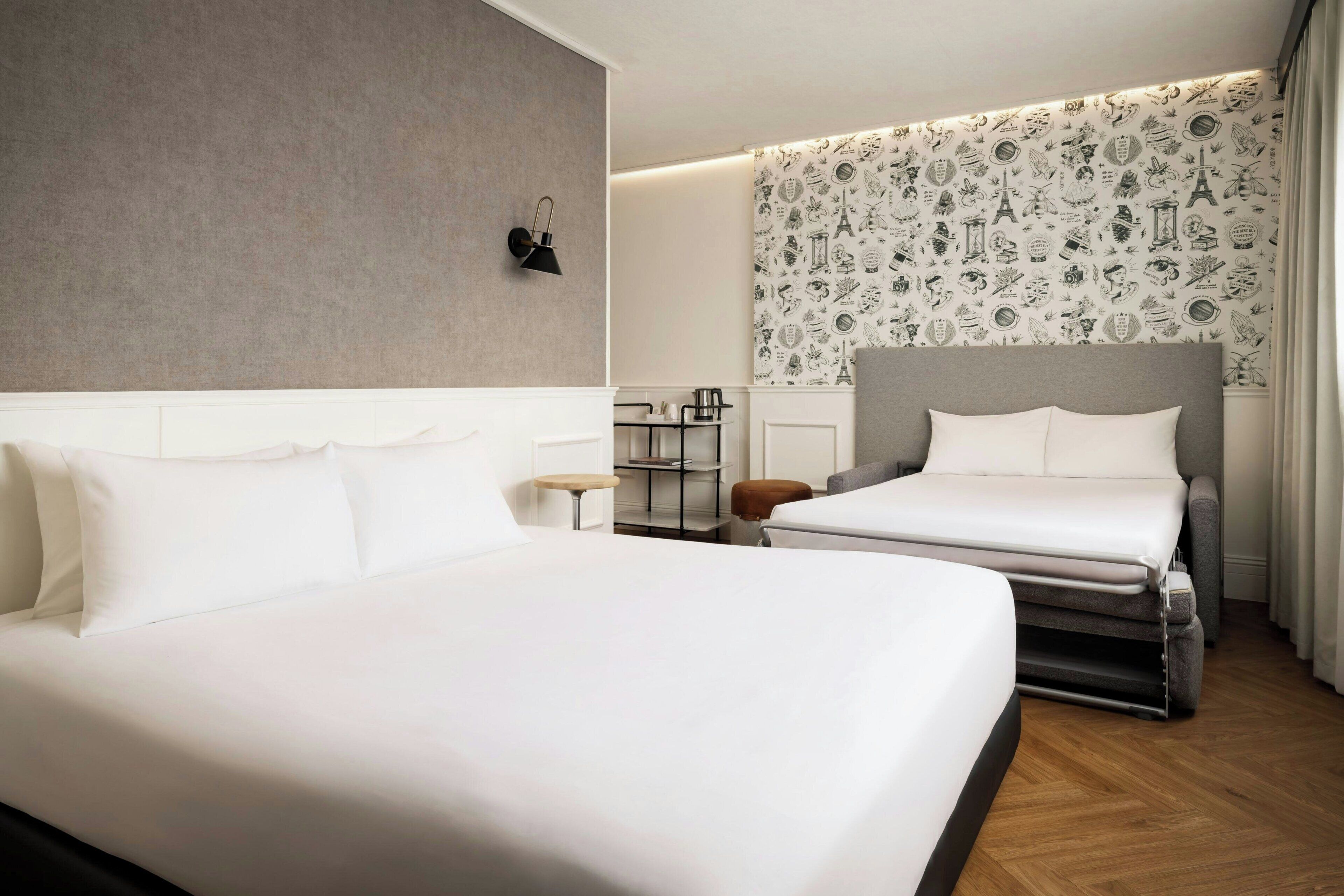 A hotel room with a large bed, a smaller bed, a bedside table, a lamp, a wall with a patterned wallpaper, and a wooden floor.