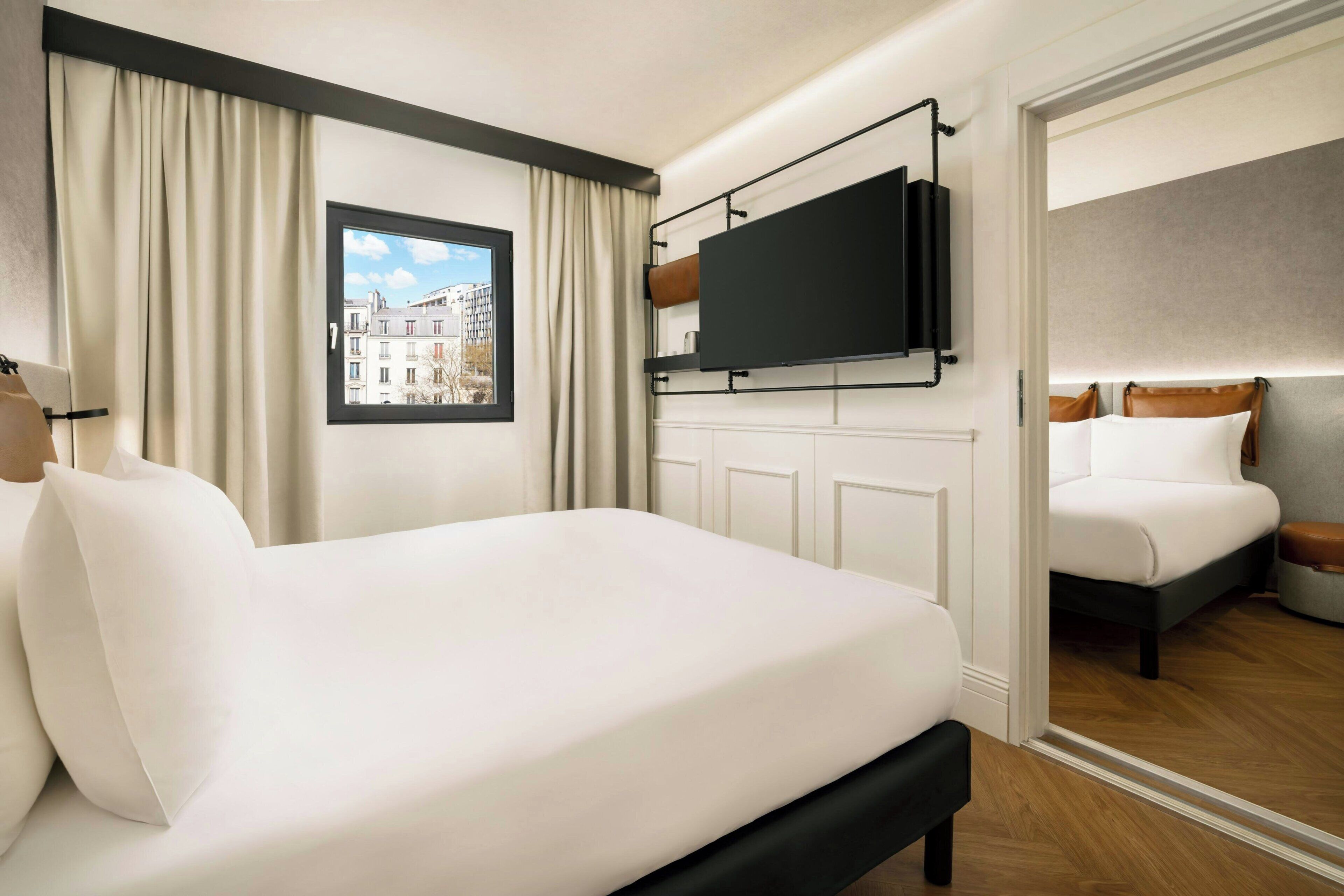 A hotel room with a bed, a TV mounted on the wall, a window with curtains, and a view of buildings outside.
