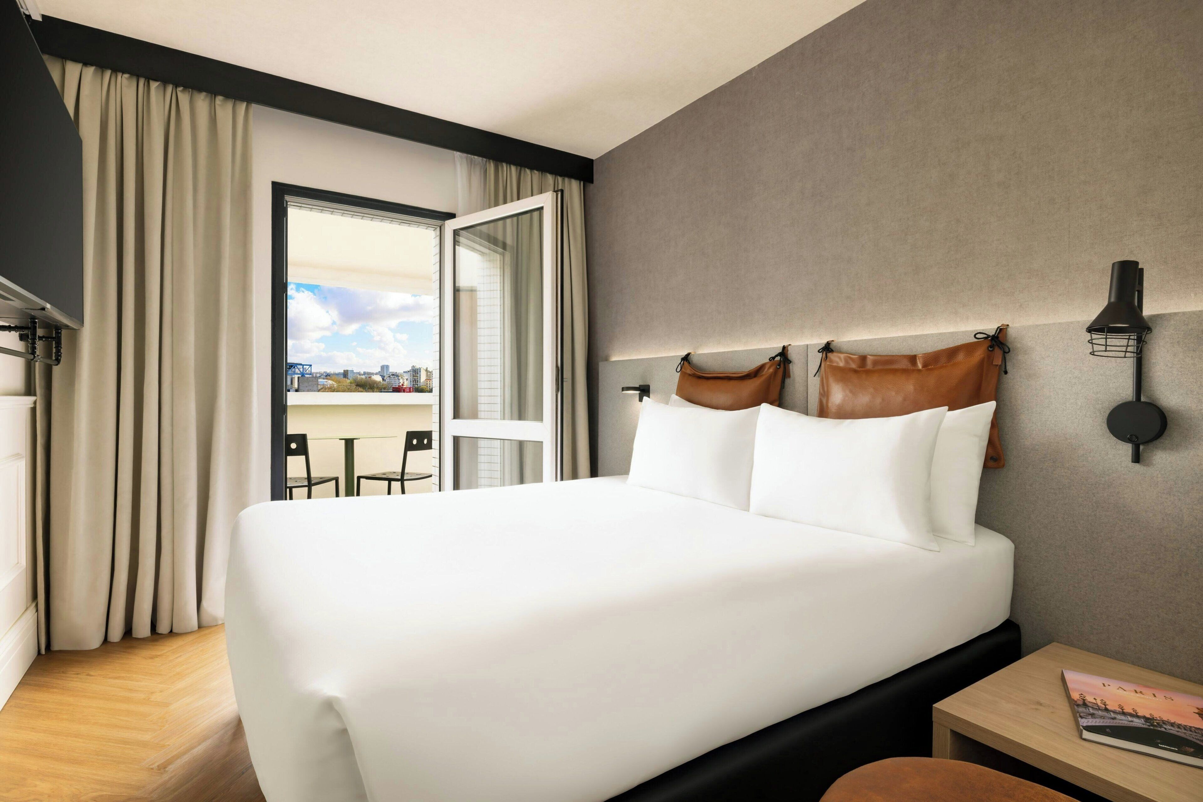 A hotel room with a large bed, a small table, a chair, a window with curtains, and a view of the city.