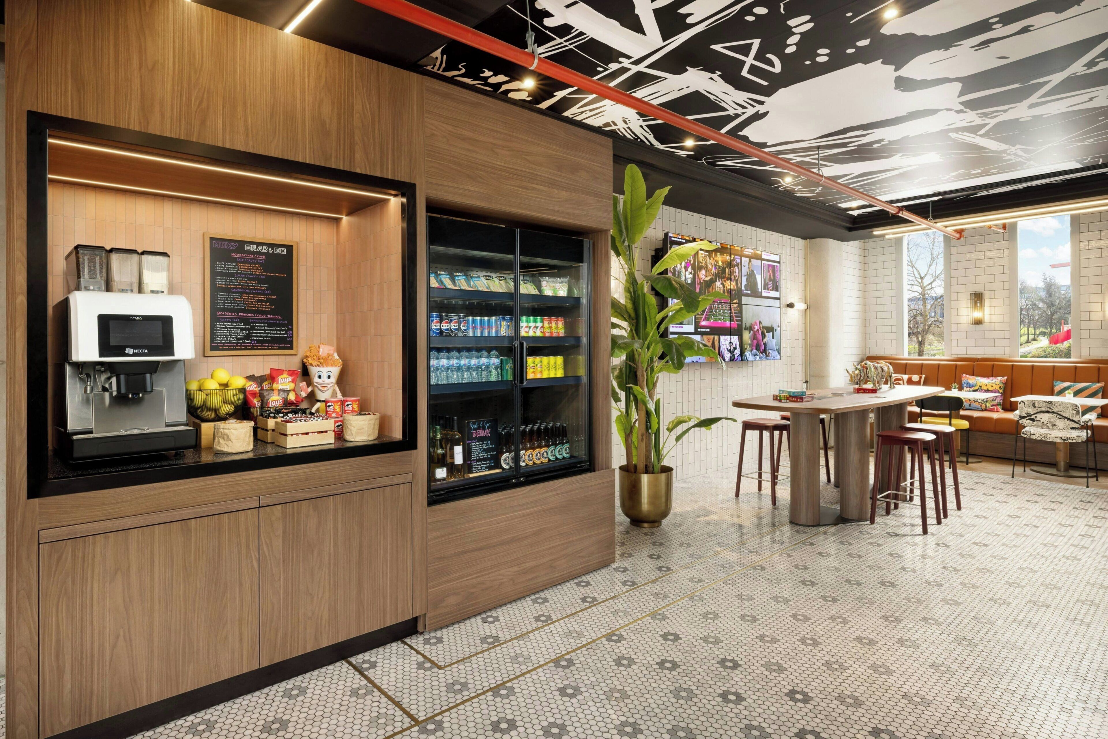 A modern cafe with a coffee machine, refrigerated drinks, and a seating area with a plant.