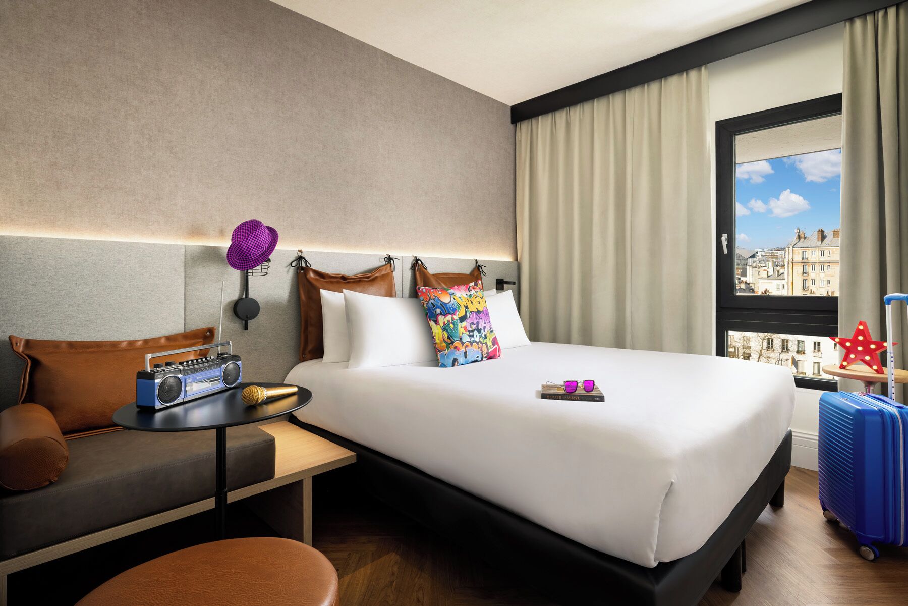 Habitació d'hotel amb un llit, un escriptori, una cadira, una maleta i una finestra que dona a una vista de la ciutat.