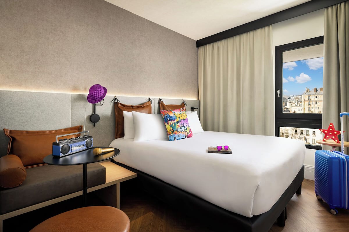 Habitación de hotel con cama, escritorio, silla, maleta y ventana con vista a la ciudad.