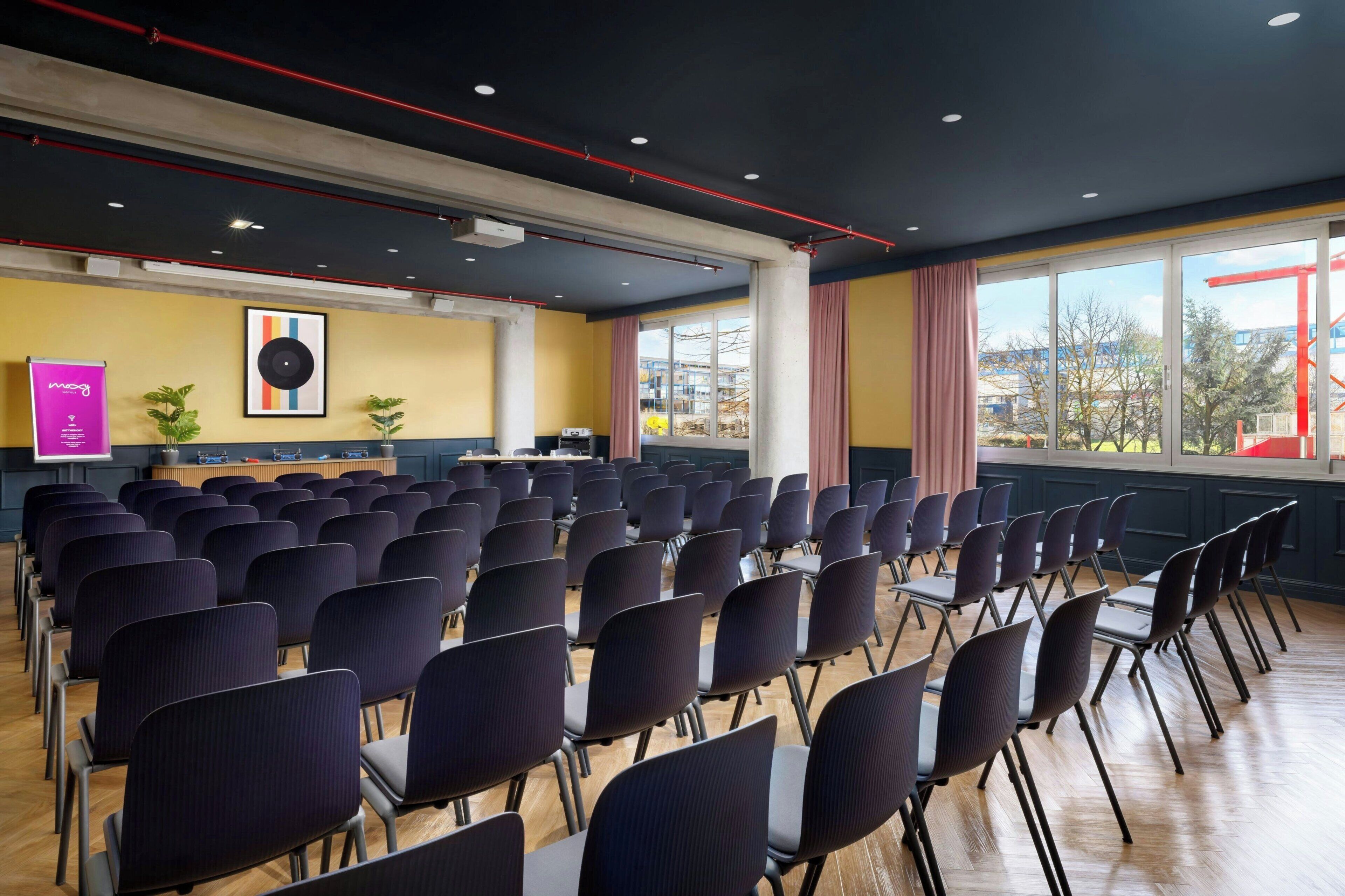 Uma sala de conferências com fileiras de cadeiras, um púlpito e uma janela grande com vista para edifícios.