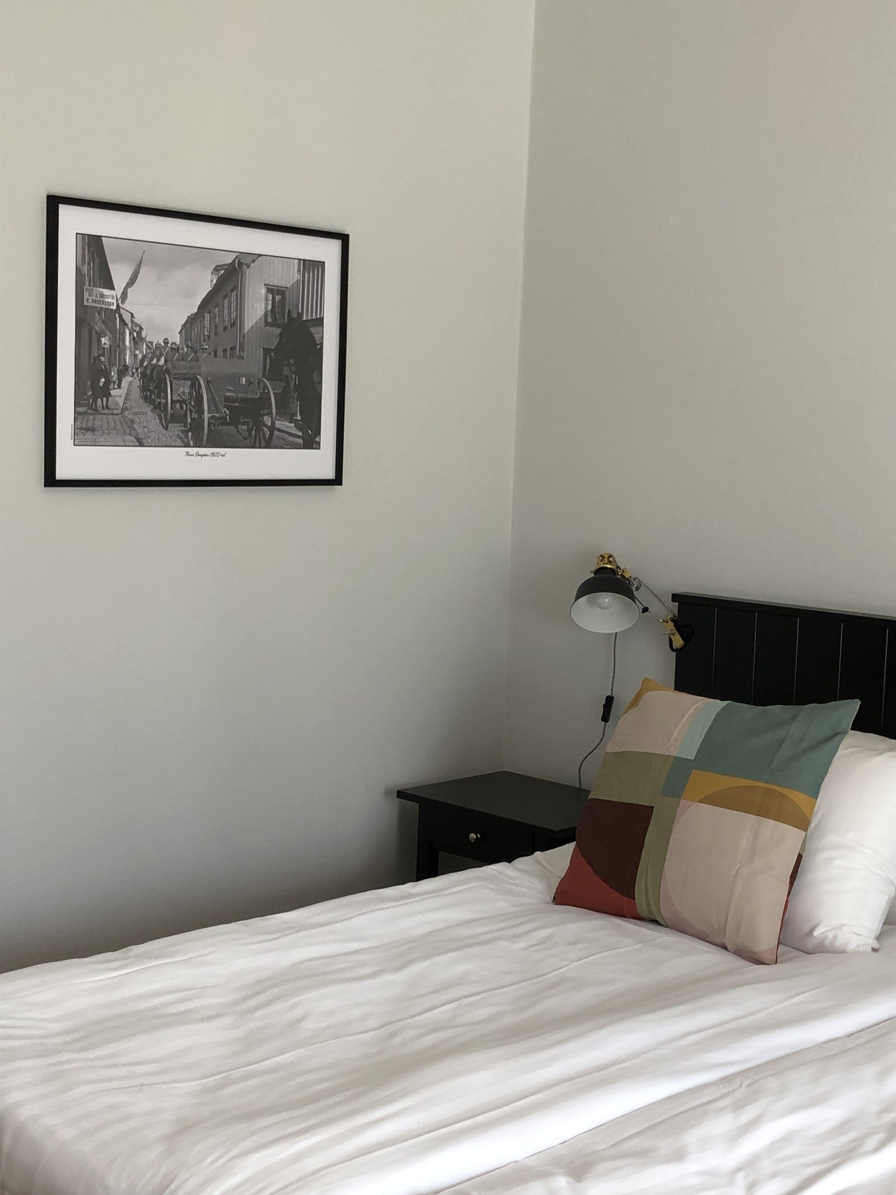 Una cama bien hecha con una colcha blanca y almohadas, una mesita de noche con una lámpara, y una fotografía en blanco y negro enmarcada en la pared.