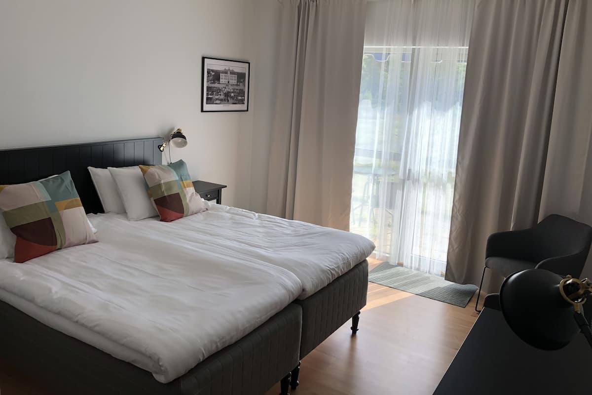A bedroom with a bed, a nightstand, a desk, a chair, a window with curtains, and a framed picture on the wall.