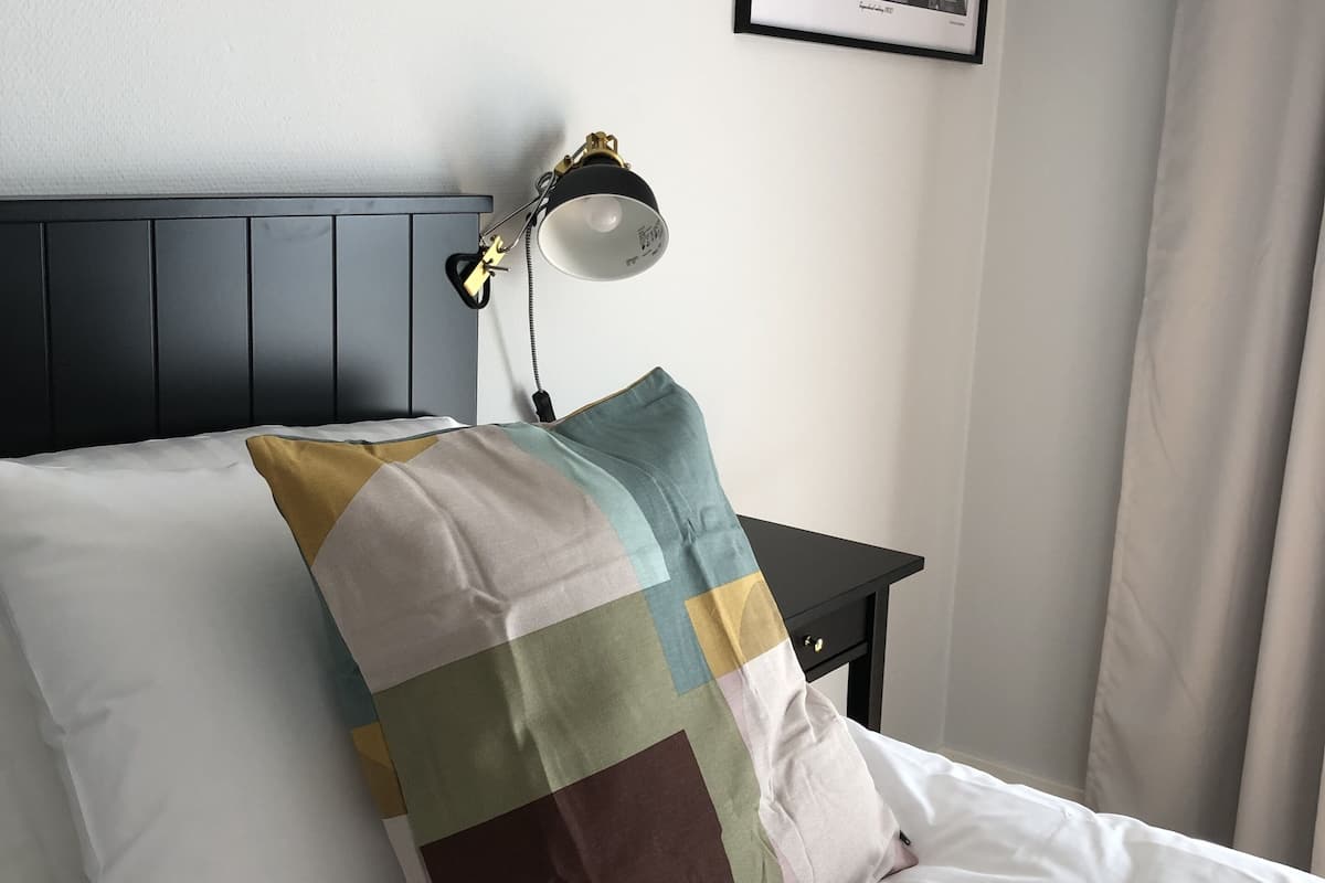 A neatly made bed with a white pillow and a colorful patchwork pillow, a black nightstand with a lamp, and a framed photo on the wall.
