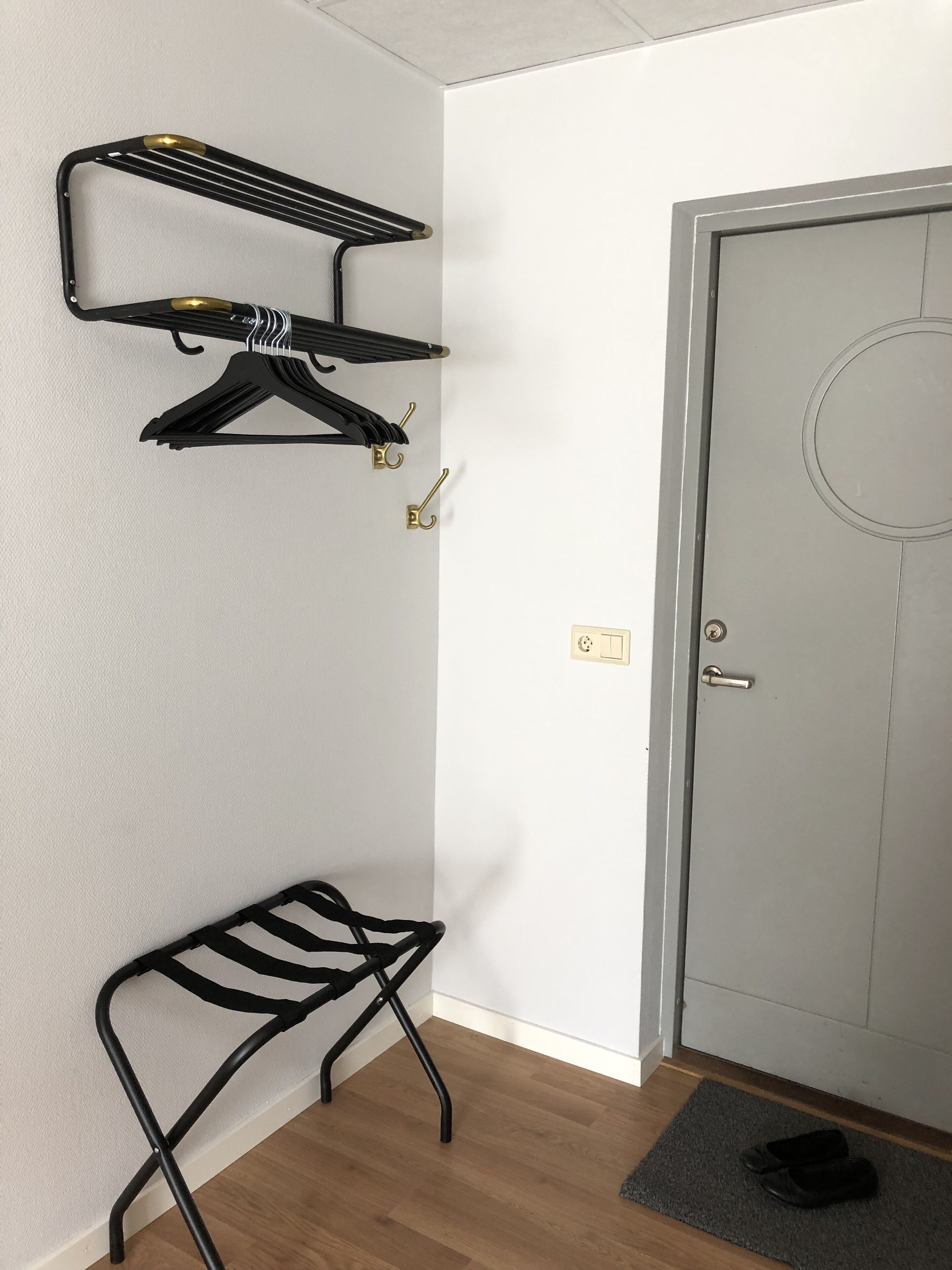 Et værelse med trægulv, en dør med et rundt vindue, en sort metalbænk og en metalreol med kroge på væggen.