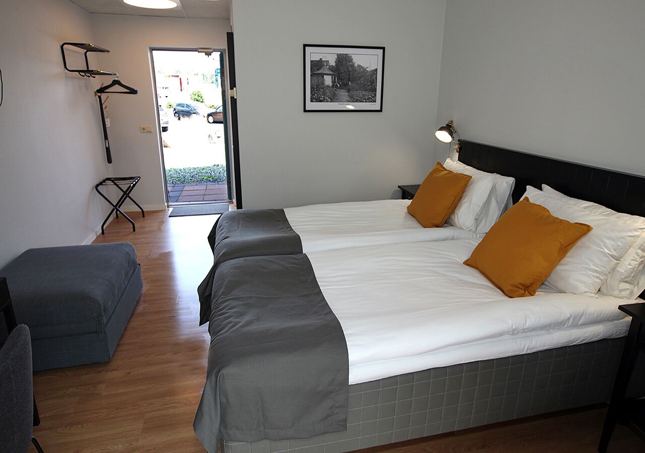 Habitación de hotel con dos camas, un banco gris, una puerta que da al exterior y un cuadro en la pared.