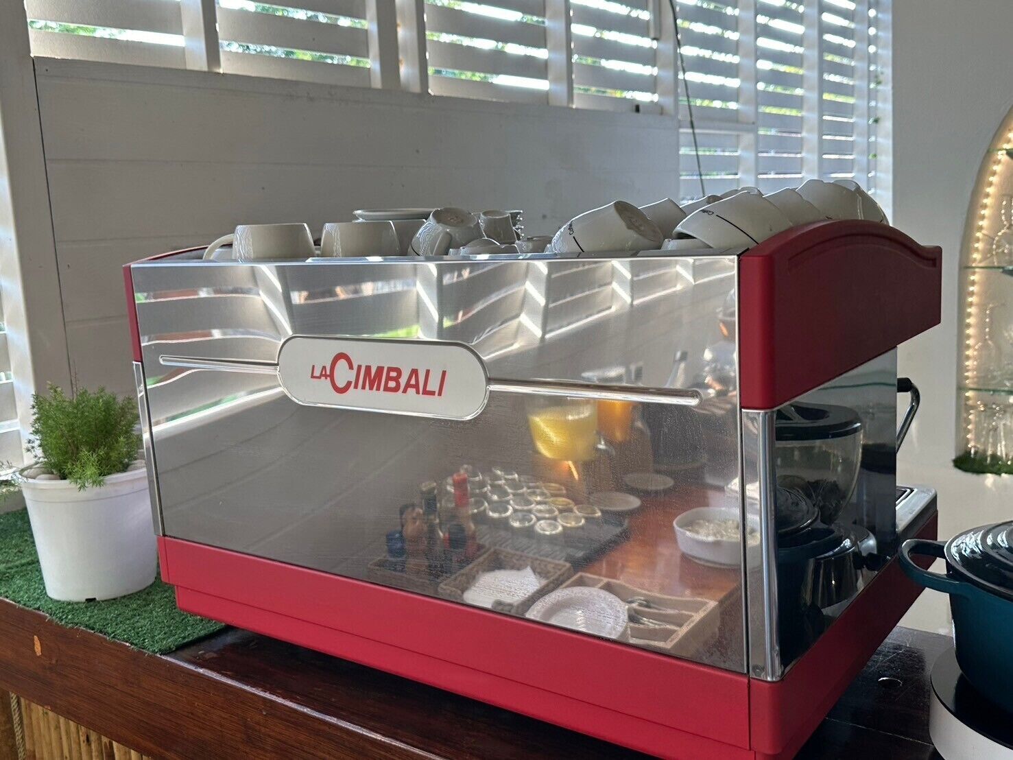 Máquina de café espresso La Cimbali con exterior de acero inoxidable y base roja, ubicada sobre una encimera con una planta en maceta cerca.
