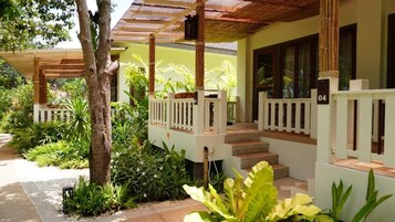 Uma área externa coberta com pérgola de madeira, degraus que levam a um alpendre branco e vegetação exuberante.