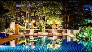 Una piscina con cubierta de madera, rodeada de árboles y mobiliario de exterior.