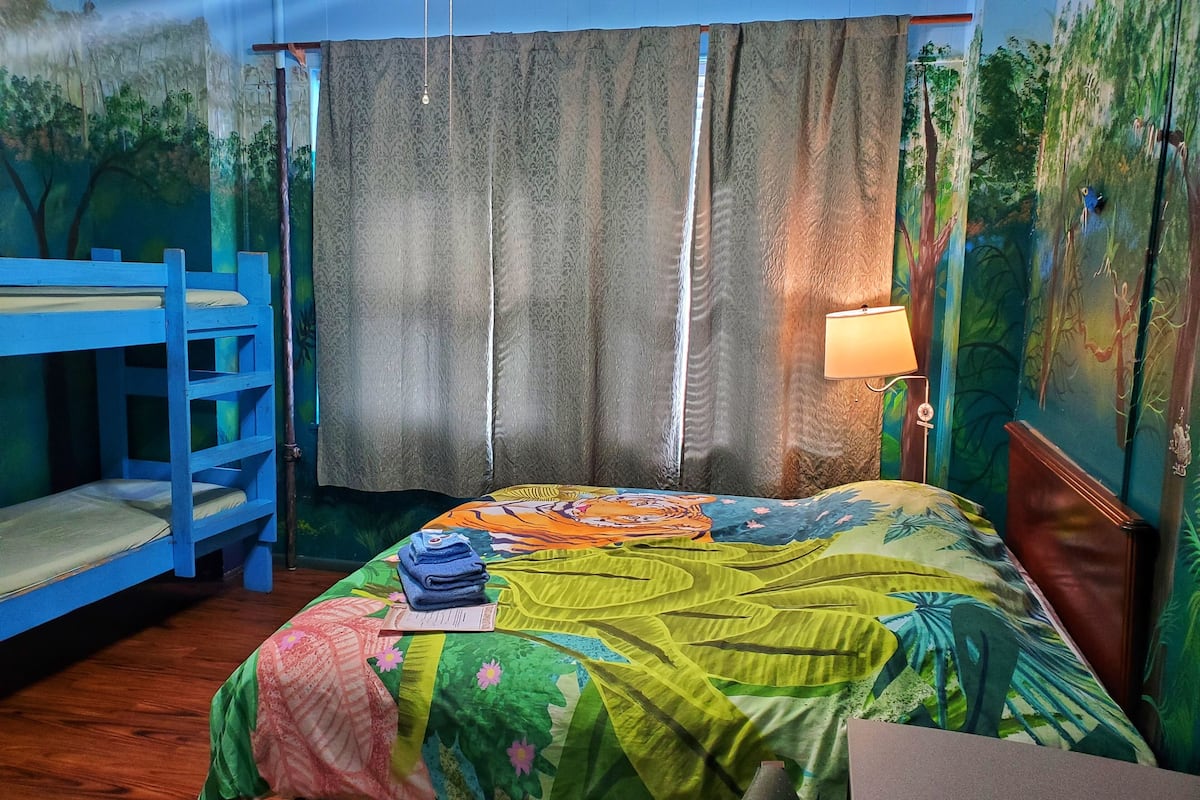 Quarto com beliches, uma cama com colcha colorida, uma luminária e um mural na parede.