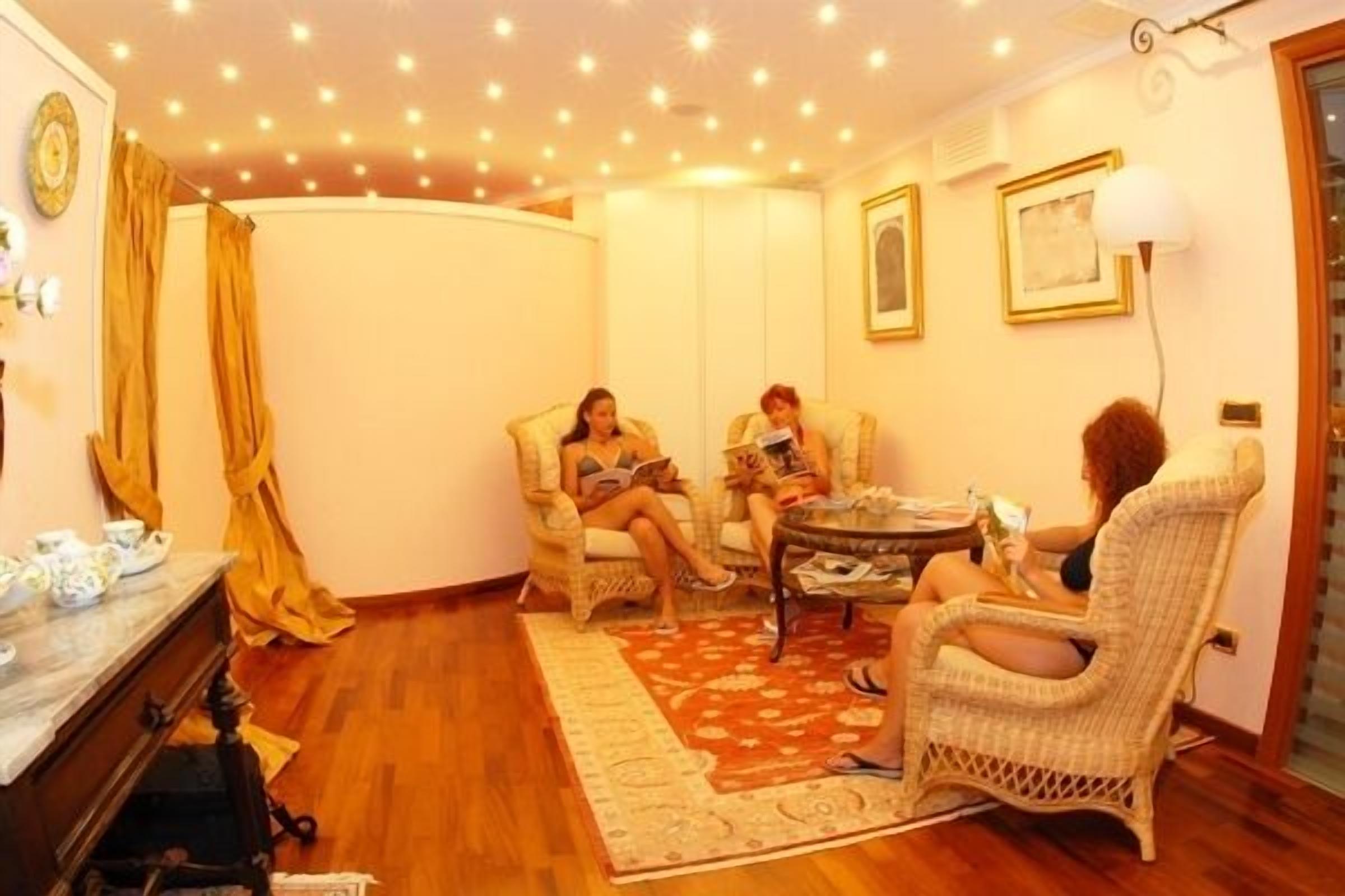 A cozy living room with a wooden floor, a patterned rug, and wicker furniture. There are two people reading, and a table with books and a lamp.
