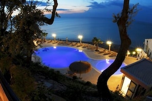 A pool area with lounge chairs and a thatched umbrella, surrounded by trees and overlooking a body of water. - Hotel Residence Tramonto (Rodi Garganico)
